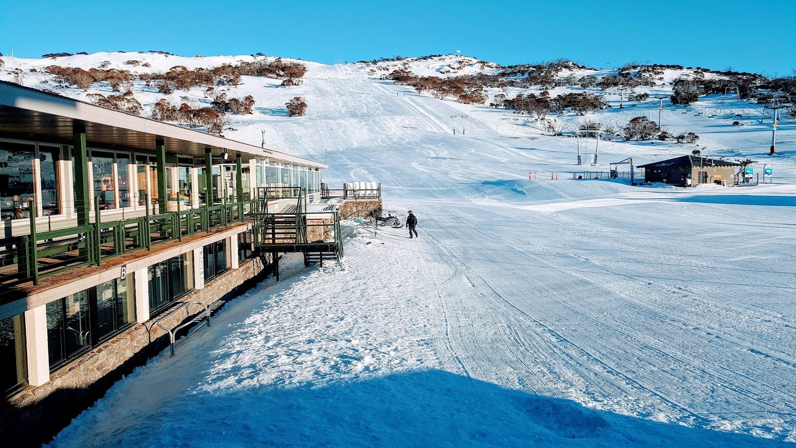 A long timber and glass lodge beside a groomed run in early light, one figure walking across the snow towards the lift