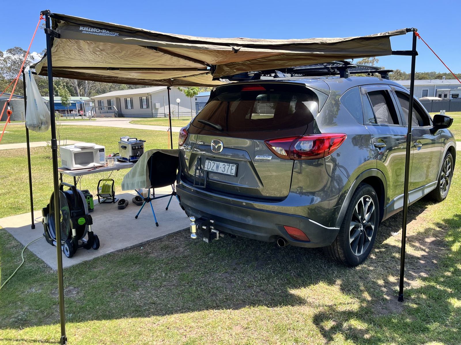 An SUV parked on grass with a large awning extended from its roof rack, camp table, chair and gear arranged underneath, cabins in the background