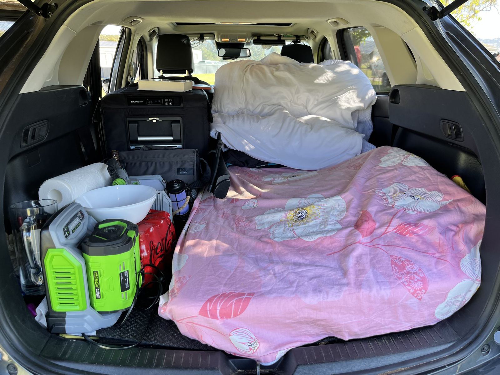 The boot of an SUV with the rear seats folded flat, made up as a bed with a mattress, pillows and a pink quilt, gear stacked at one side
