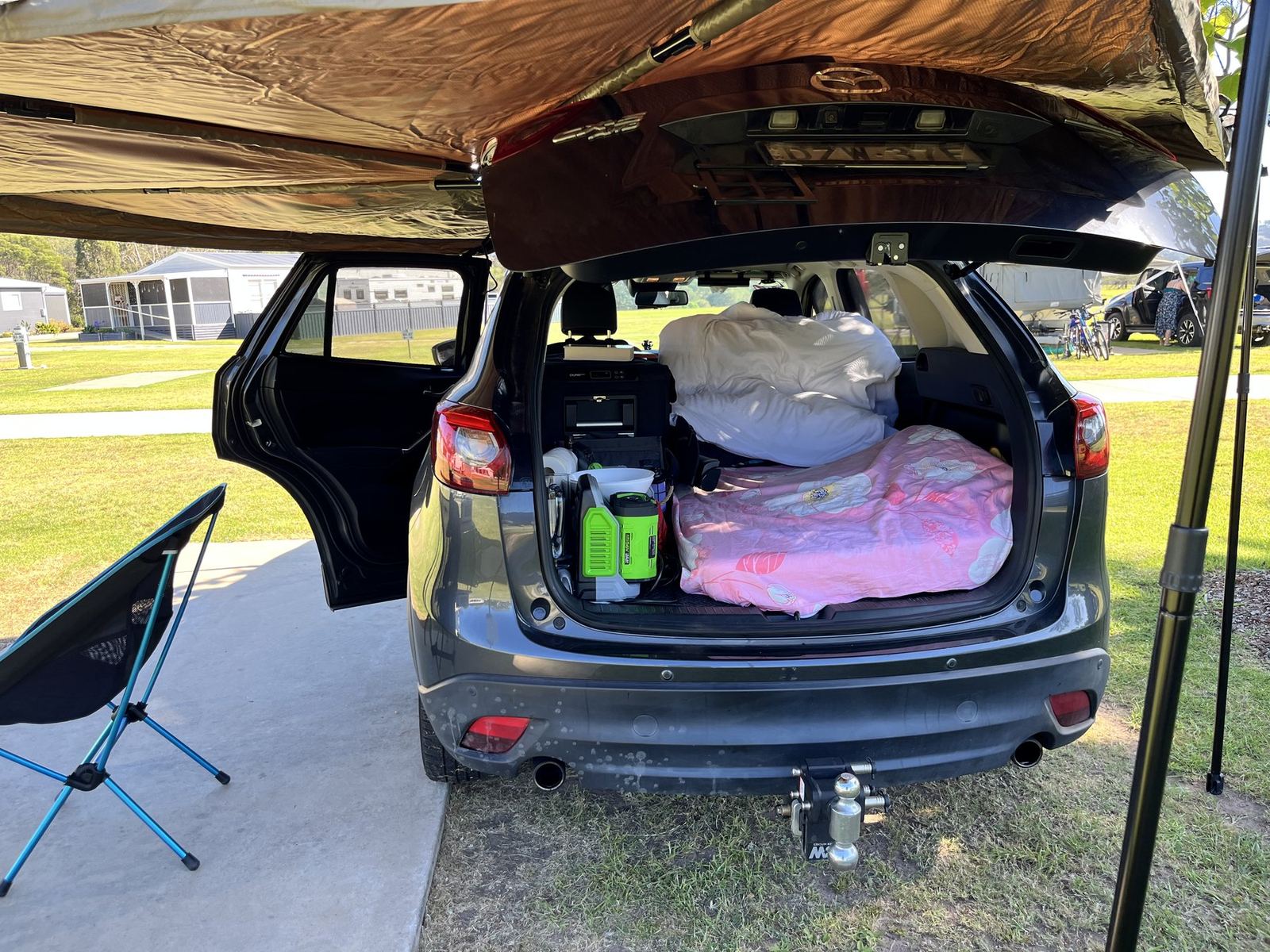 The car's tailgate open under the awning, bedding visible inside, a camp chair and battery on the grass alongside