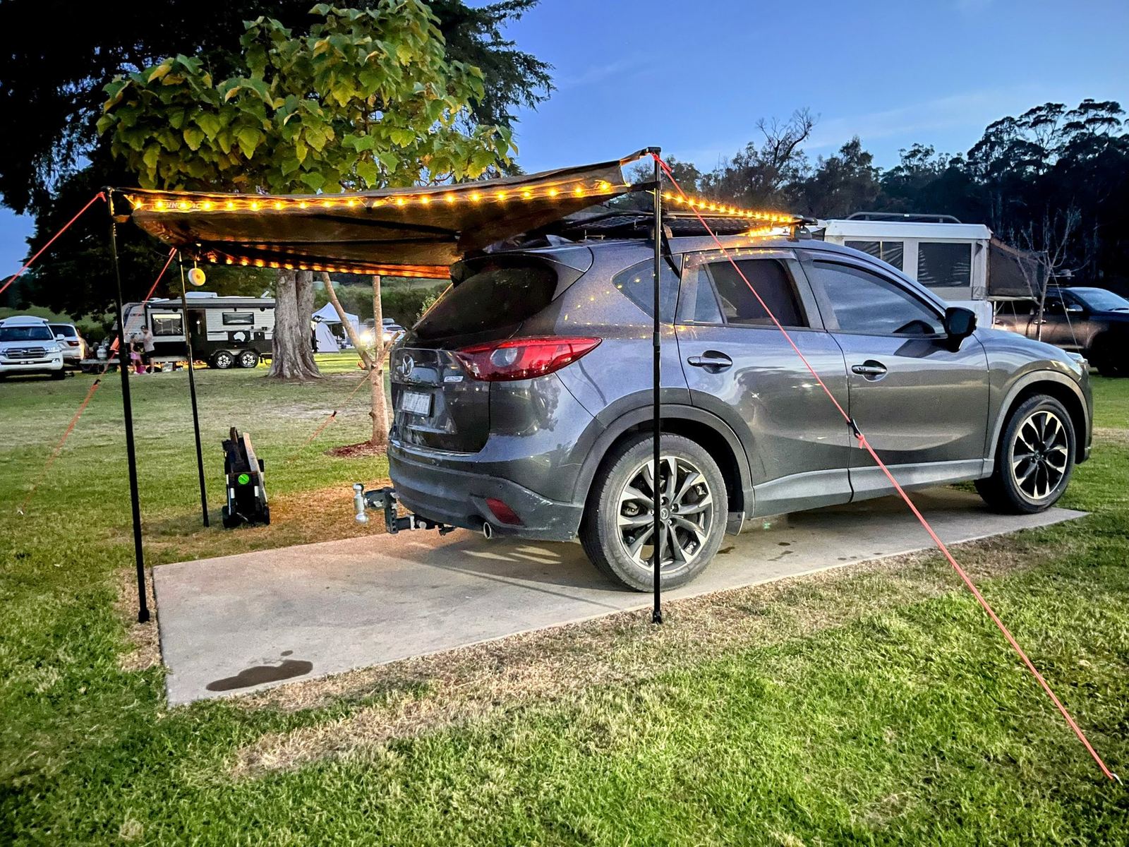 The car and awning at dusk with festoon lights strung along the awning edge, camp kitchen set up underneath
