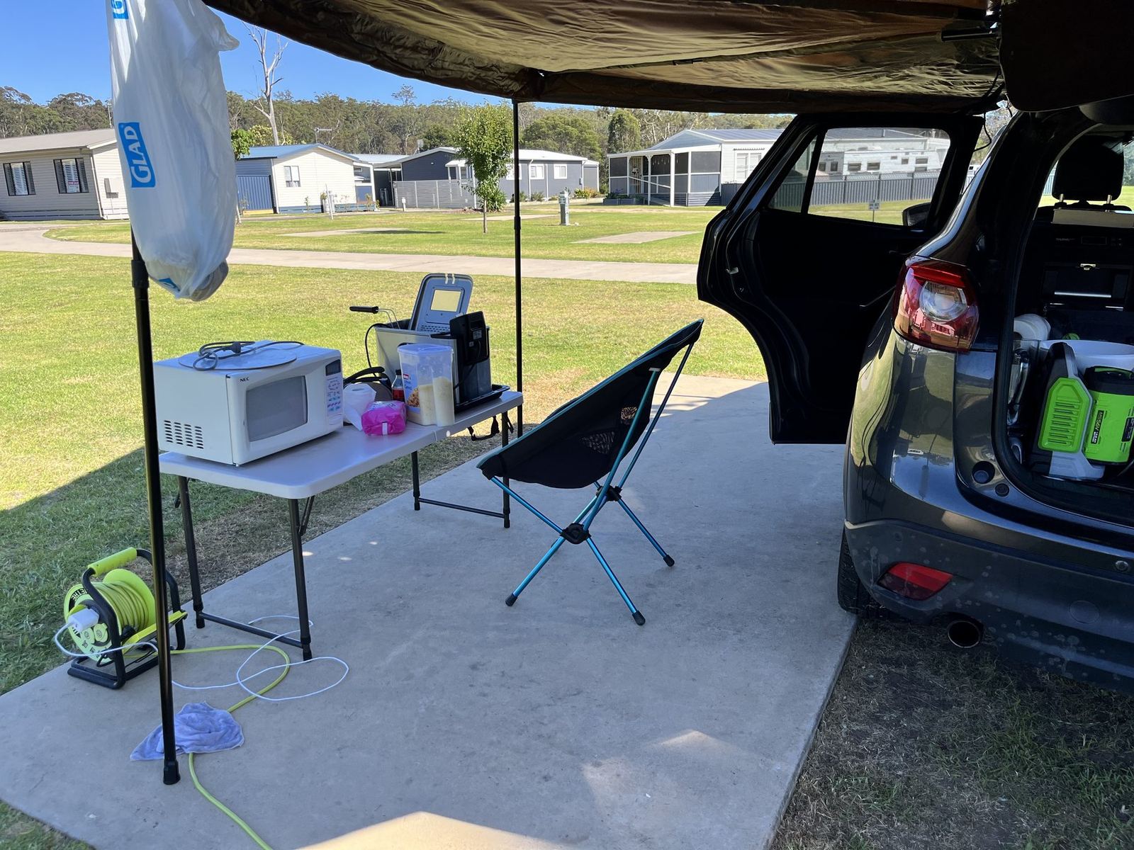 A folding table under the awning holding a microwave and other appliances, with a portable battery on the grass beneath