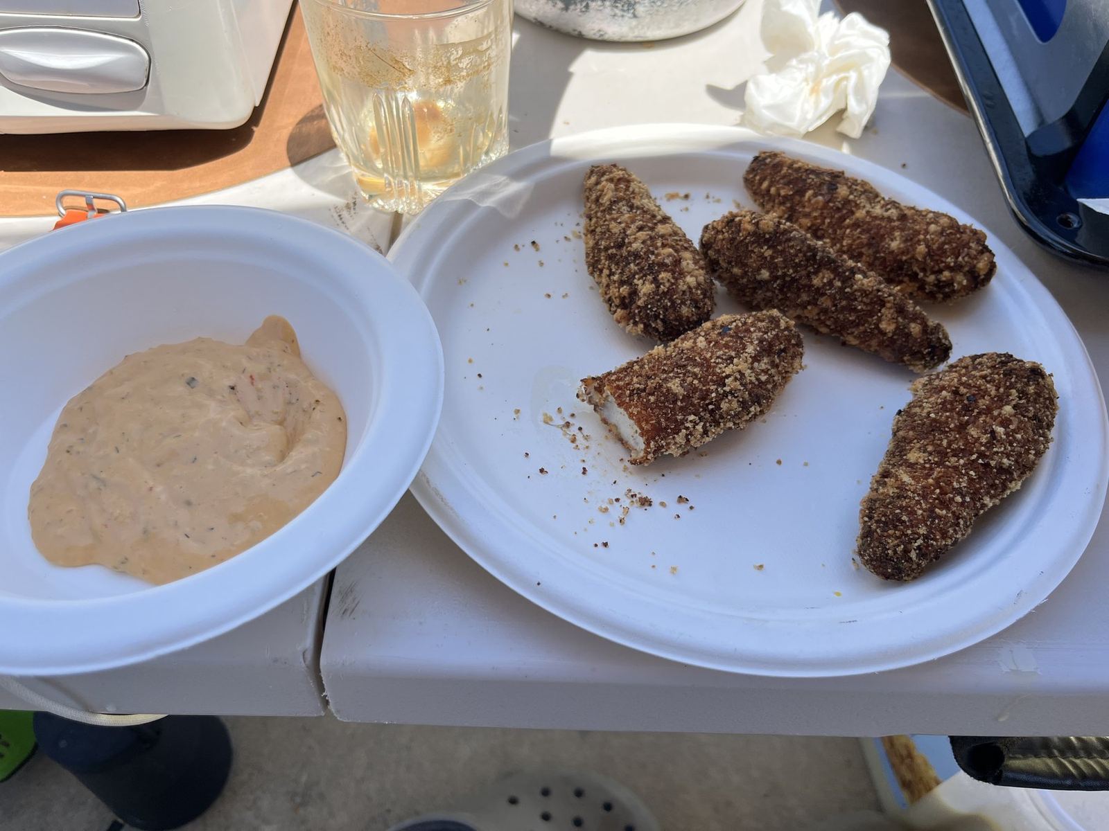 A paper plate with three crumbed cutlets and a bowl of dip on a camp table