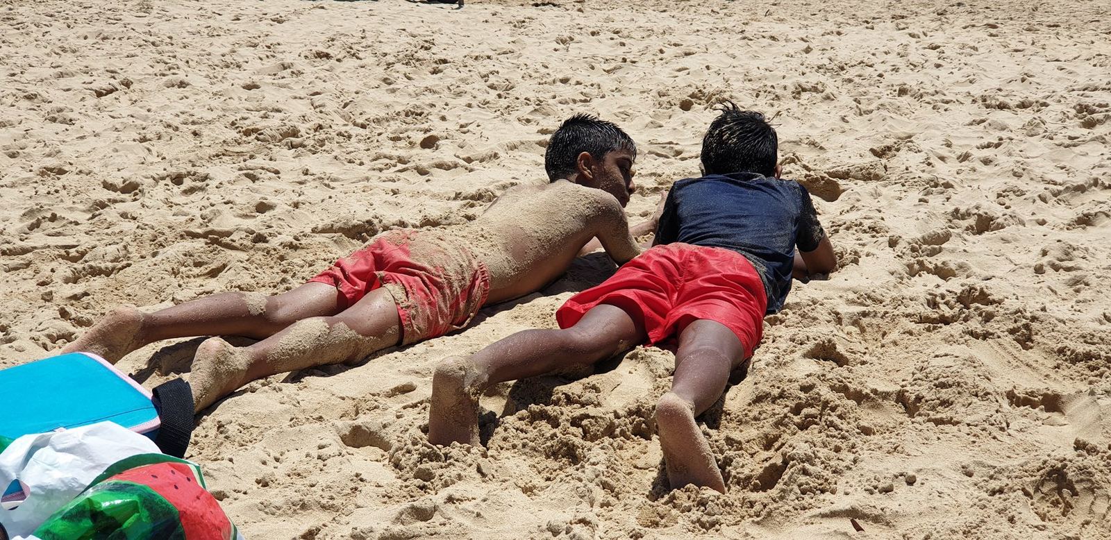 Two boys lying flat on wet sand at the edge of the water, half buried, towels and a bag on the sand beside them