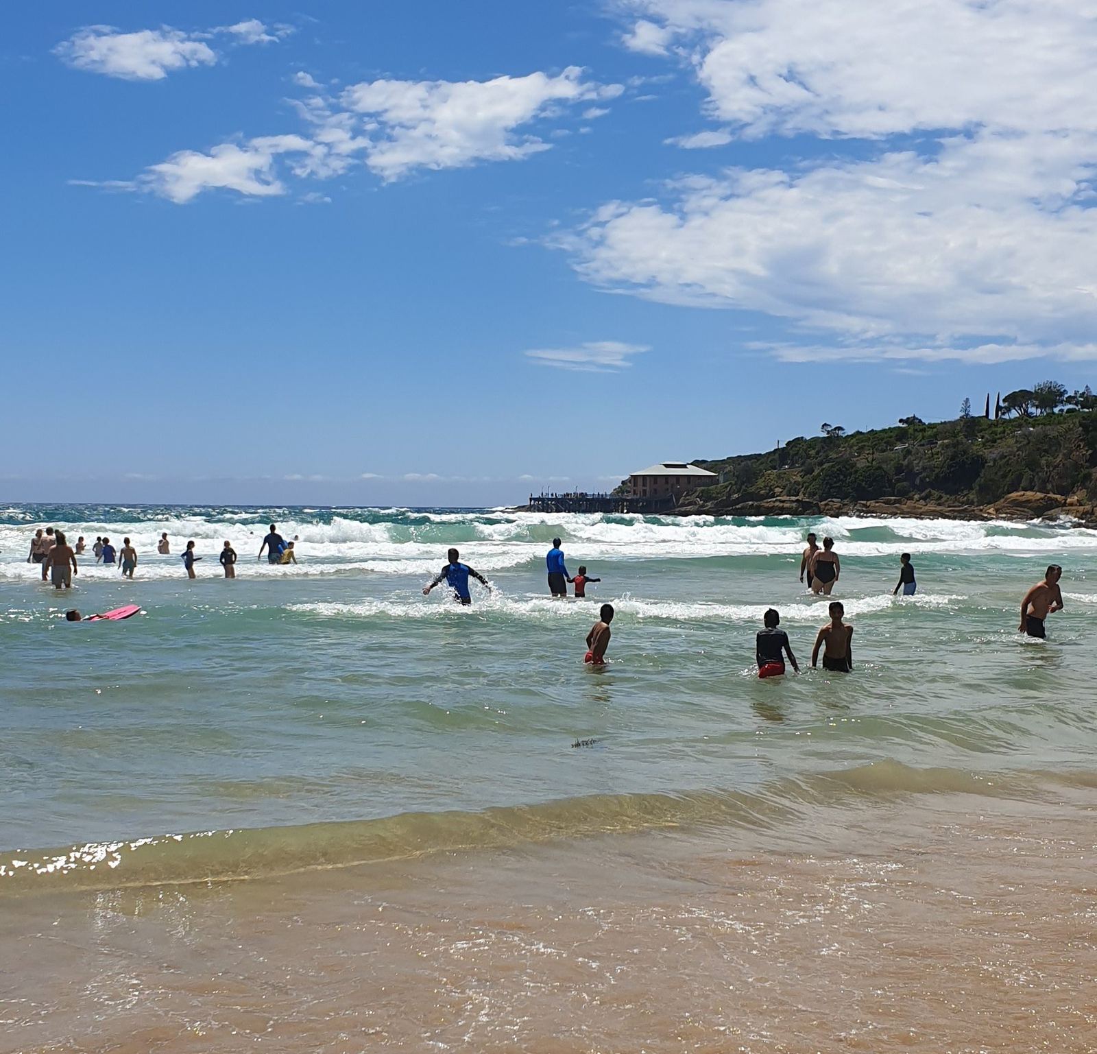 Swimmers and body-boarders in shallow green surf at a wide beach, a low wooded headland with houses on it at the far end