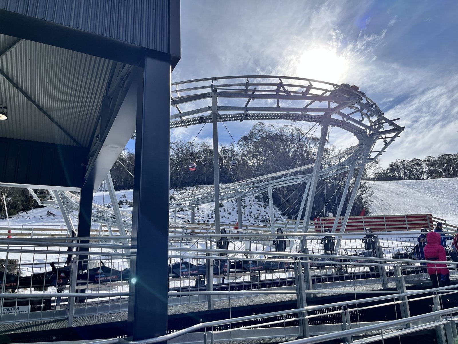 The curving steel track of the Thredbo Alpine Coaster above its loading station, snow on the slopes and gondola cabins crossing behind, under a bright sun