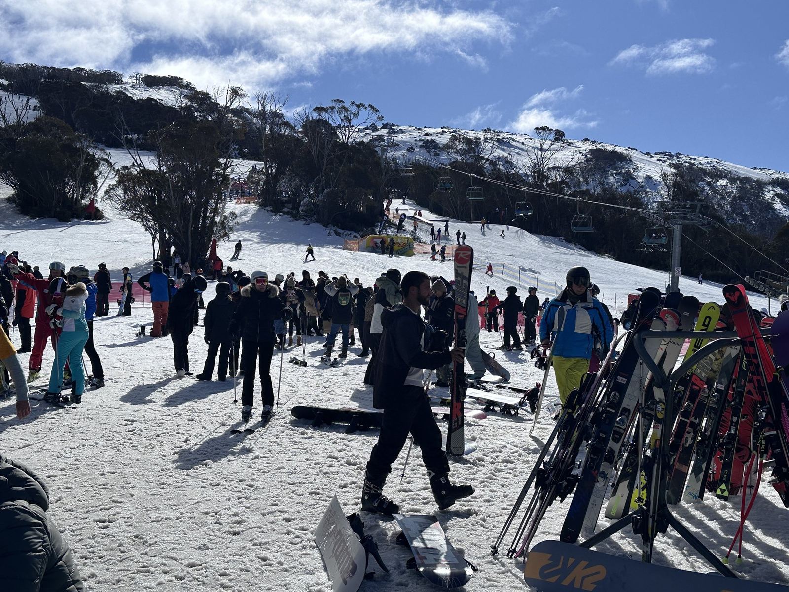 The crowded beginner slope at Thredbo in bright sun, skiers and boarders spread across thin snow with a chairlift running up the ridge behind