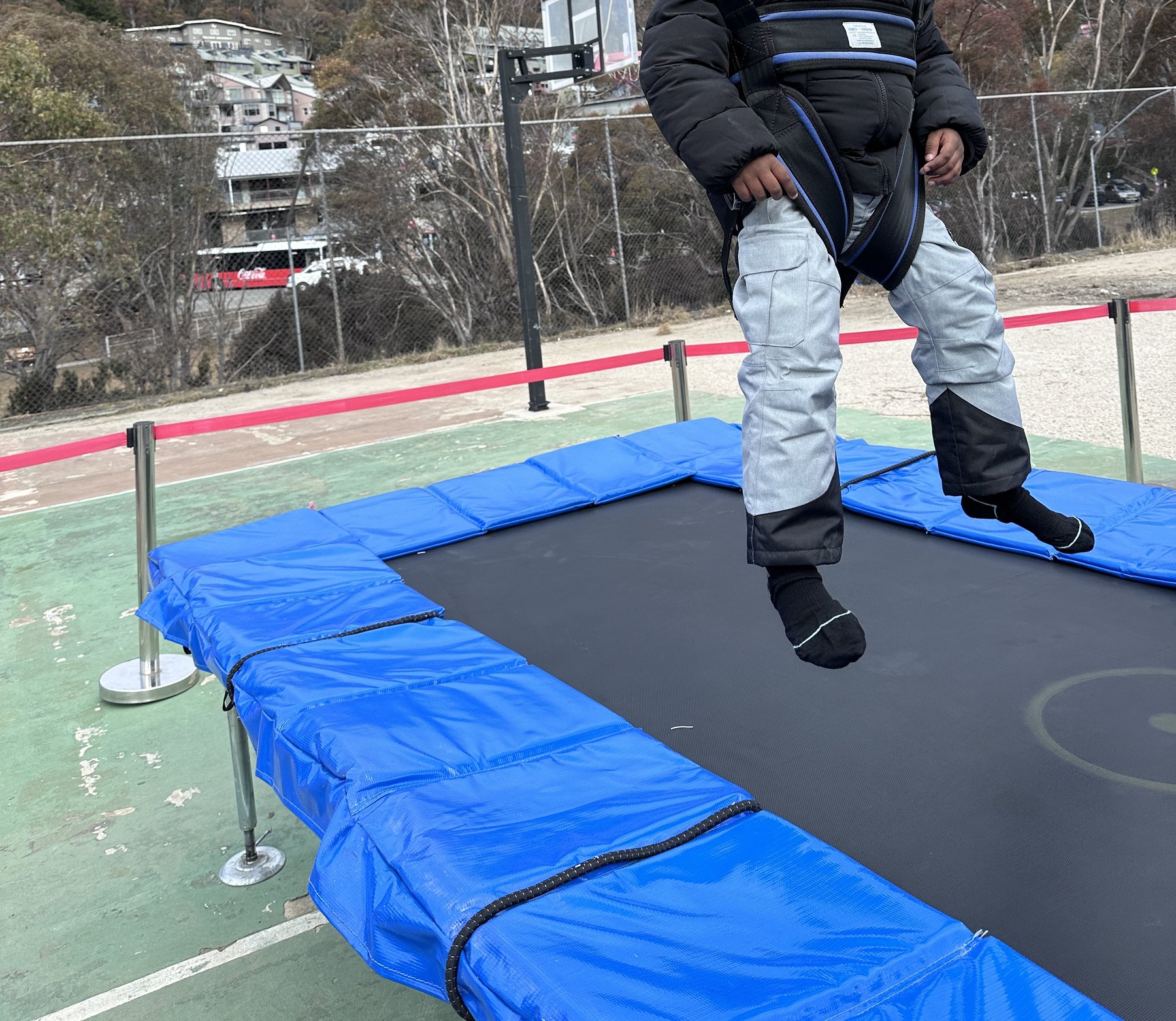 A child in snow pants and a harness bouncing on a blue-padded bungee trampoline on a fenced court, lodges on the hillside behind