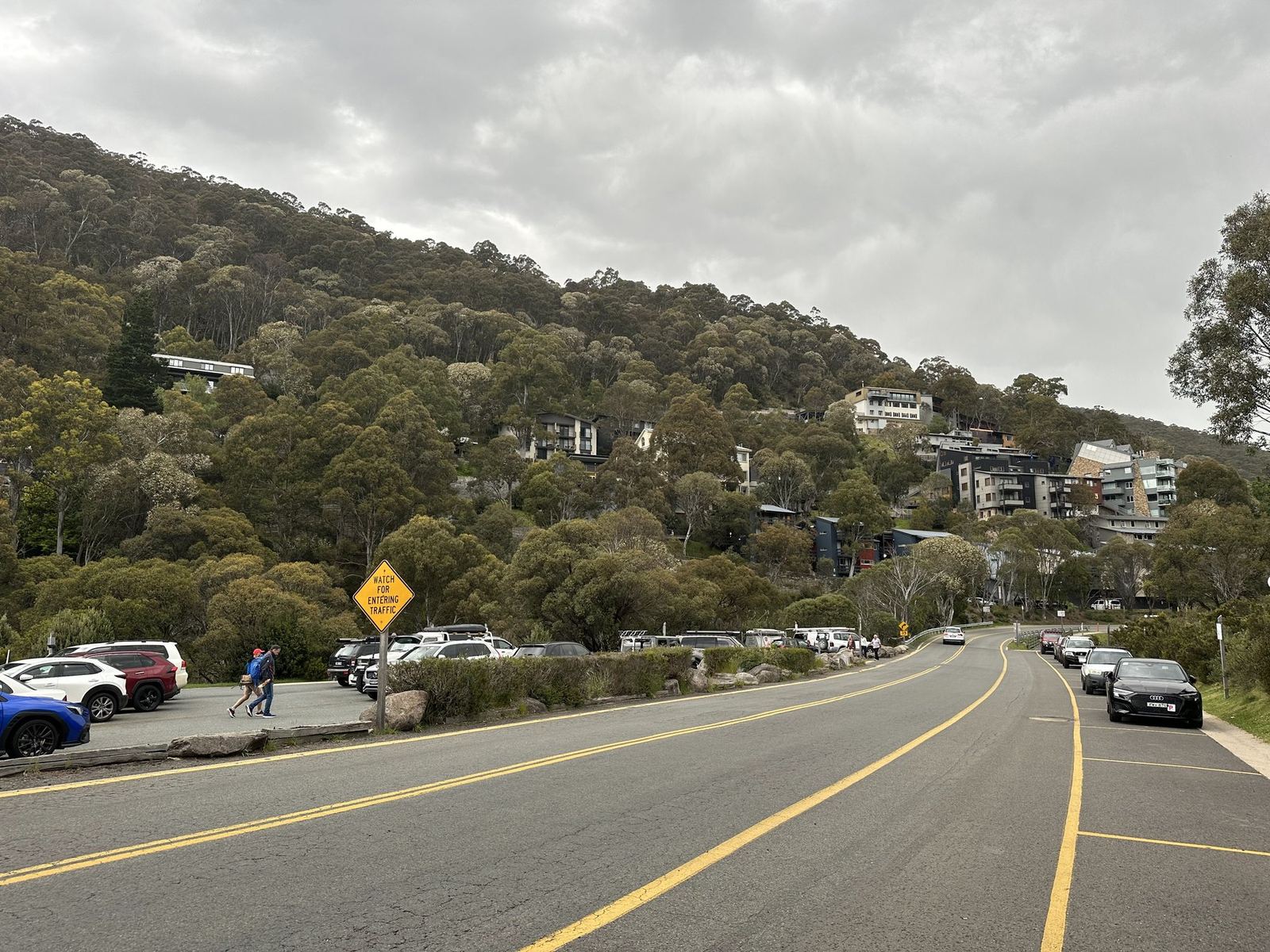 The Alpine Way curving past Thredbo village, lodges stepped up a forested hillside under heavy grey cloud
