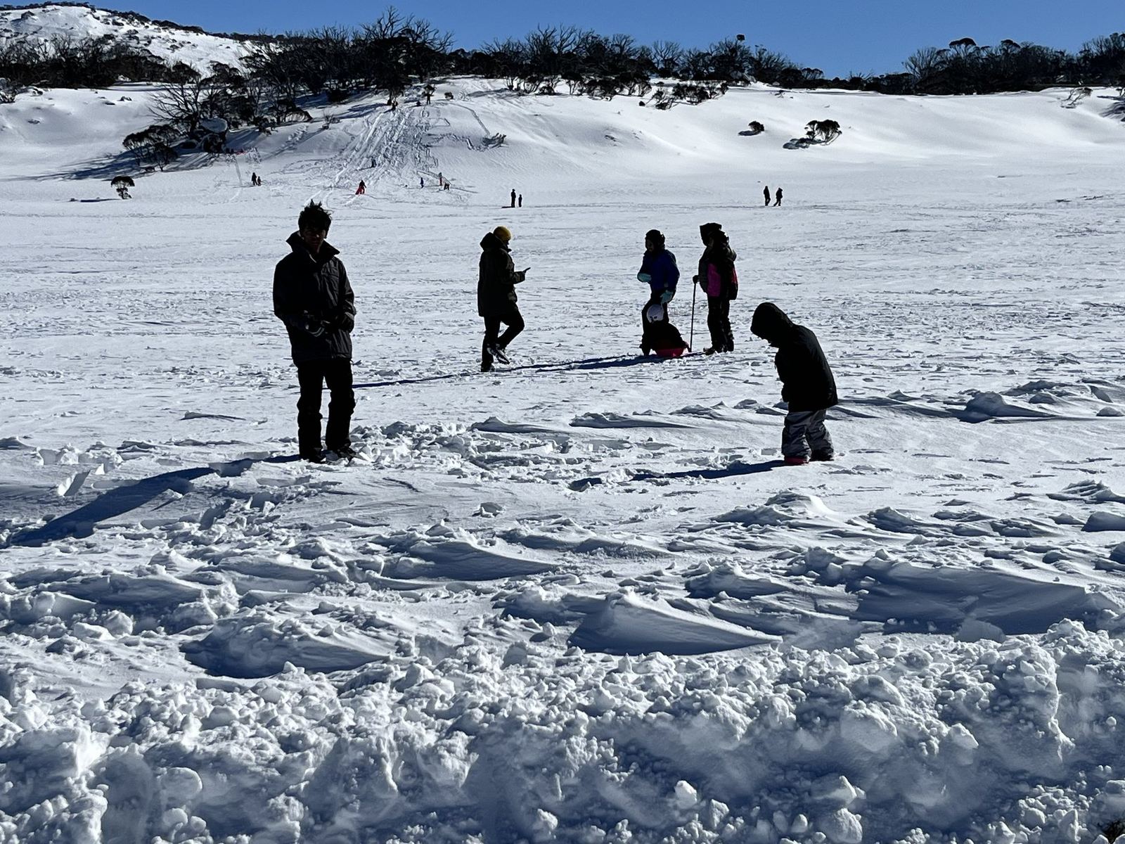 Figures silhouetted against bright snow on a toboggan slope, one kneeling beside a sled, churned snow in the foreground