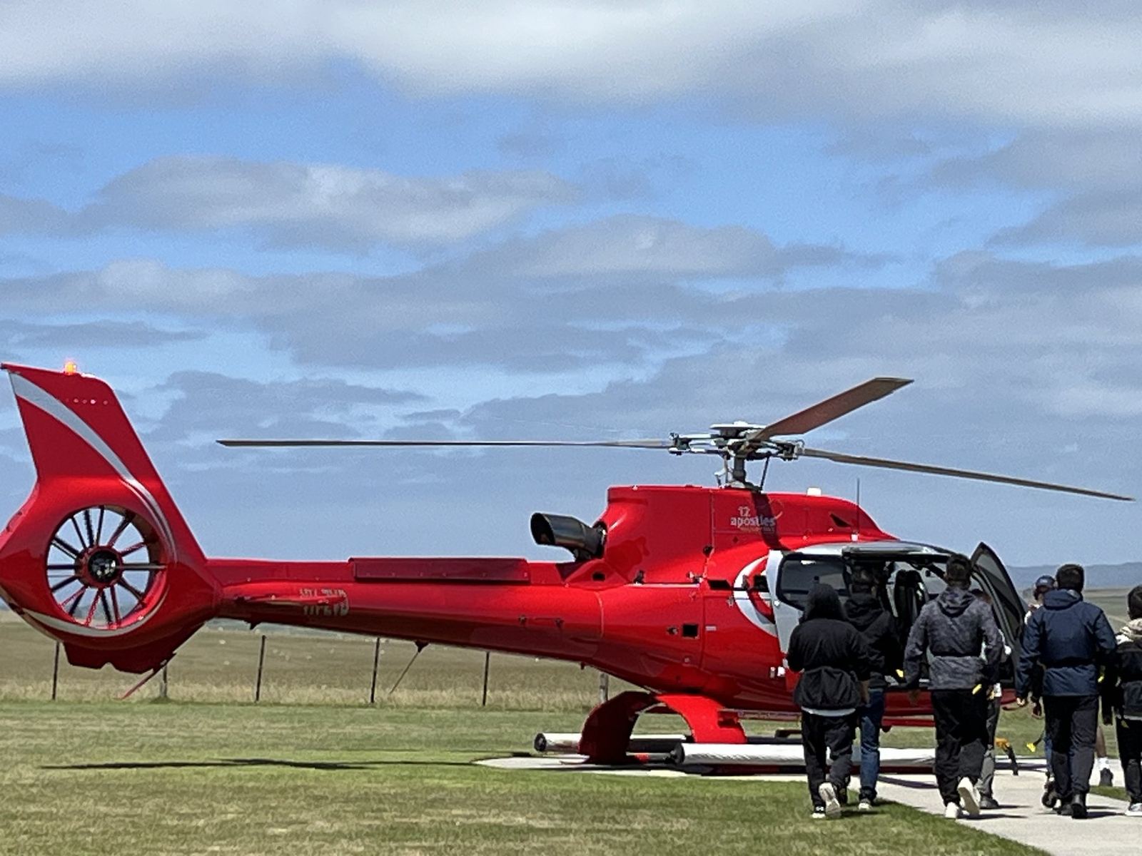 A red helicopter on the grass with its door open and a group walking towards it across the field
