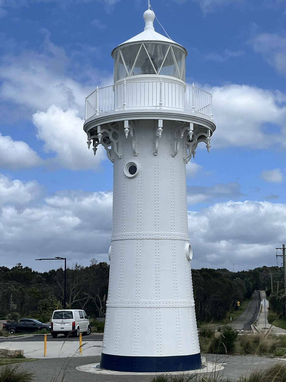 The lighthouse from its base looking up, white iron tower with a railed gallery and lantern room against blue sky and cloud