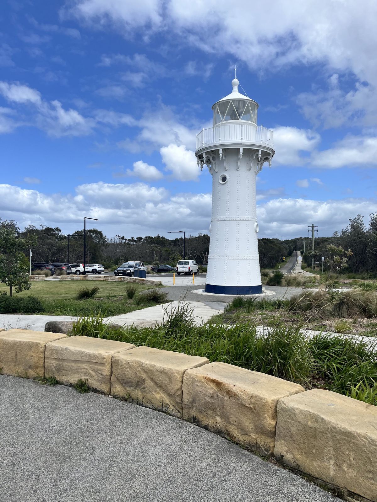 A small white lighthouse standing on a grassy headland, a sandstone wall curving along the path in the foreground and cars parked beyond