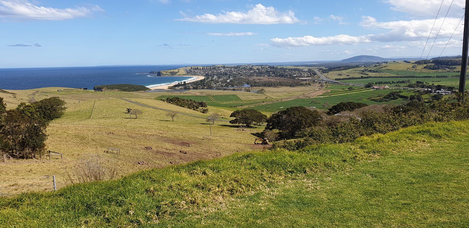 A wider view from the same hill, dairy paddocks running down to the coast with the town, the highway and distant hills beyond