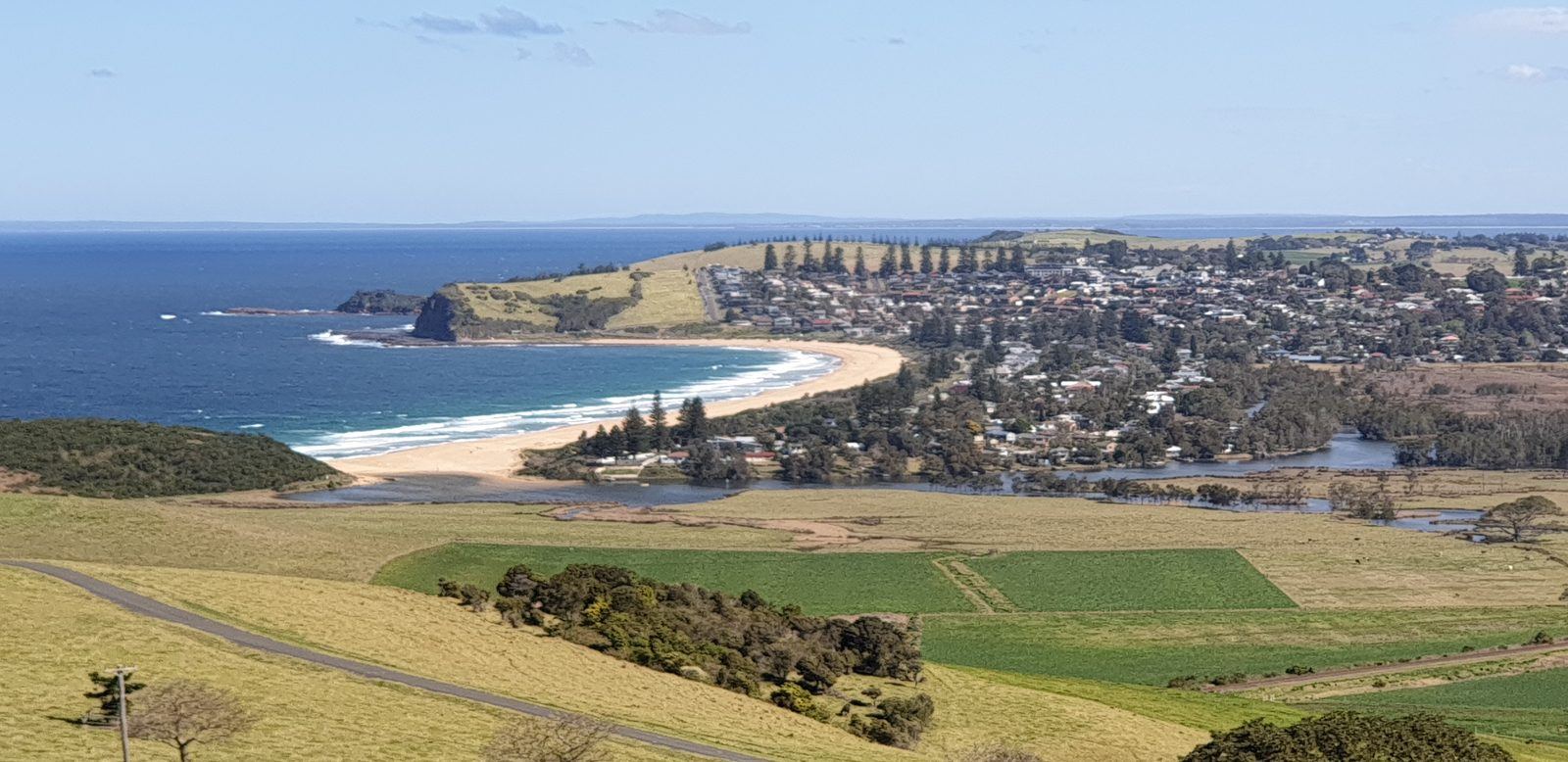 A high view over a long curved surf beach with a lagoon behind it, a small coastal town and green dairy farmland running down to the sea