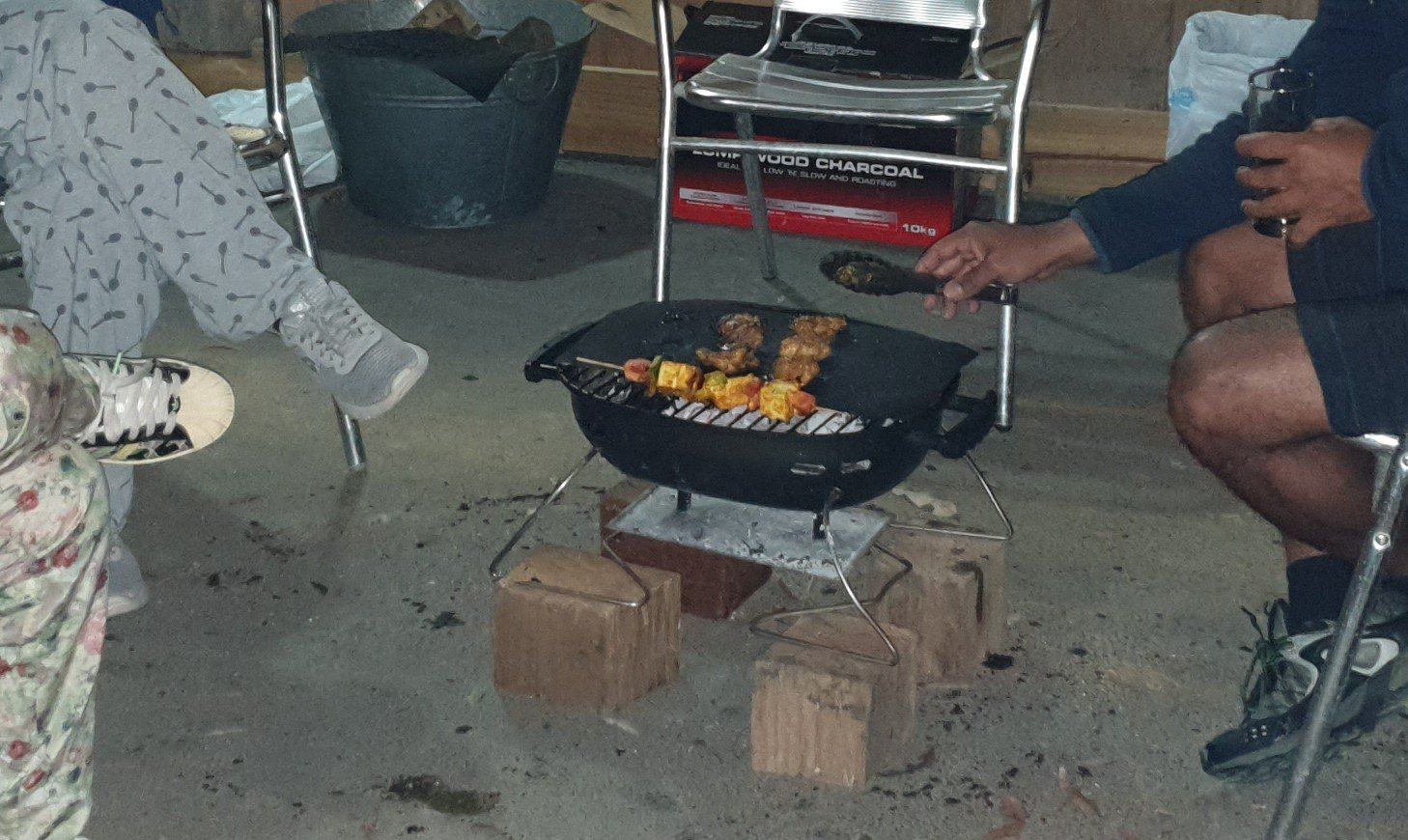 A hand reaching over a charcoal barbecue to turn skewers, people seated on either side in camp chairs on a concrete slab at night