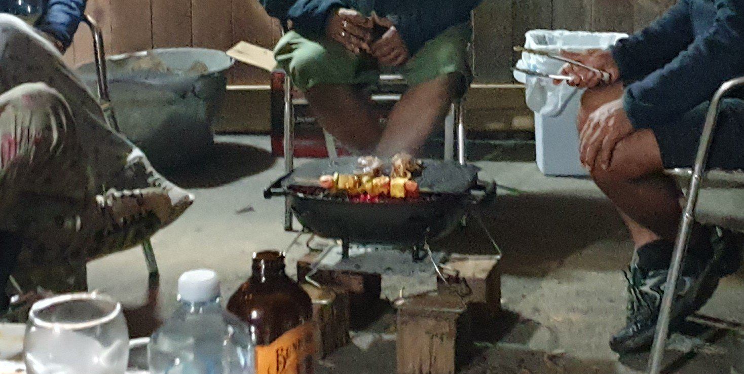 People sitting in camp chairs in a semicircle around a small charcoal barbecue on concrete outside a cabin at night