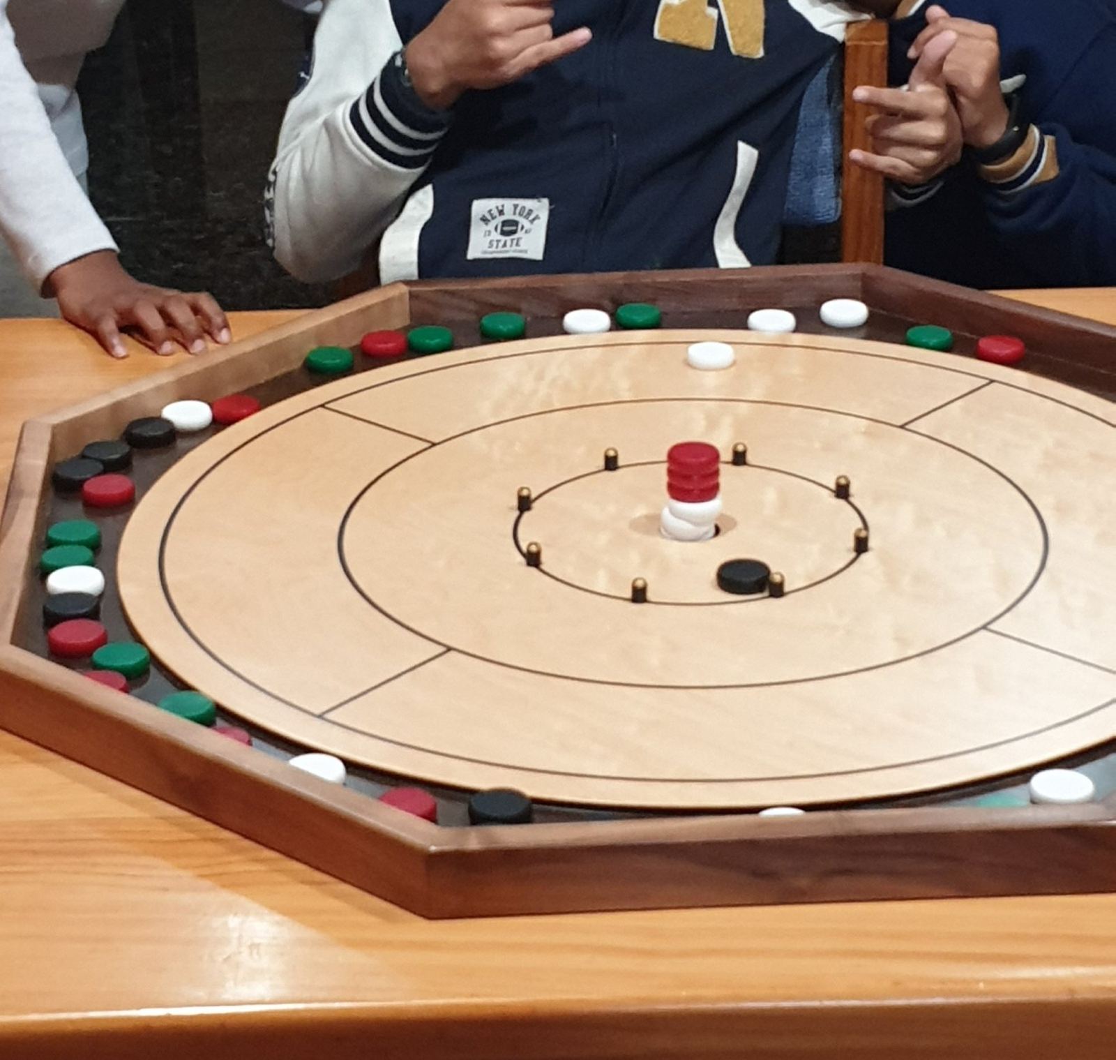 A wooden octagonal crokinole board on a table mid-game, red white green and black discs around the rim, two players' hands at the edge