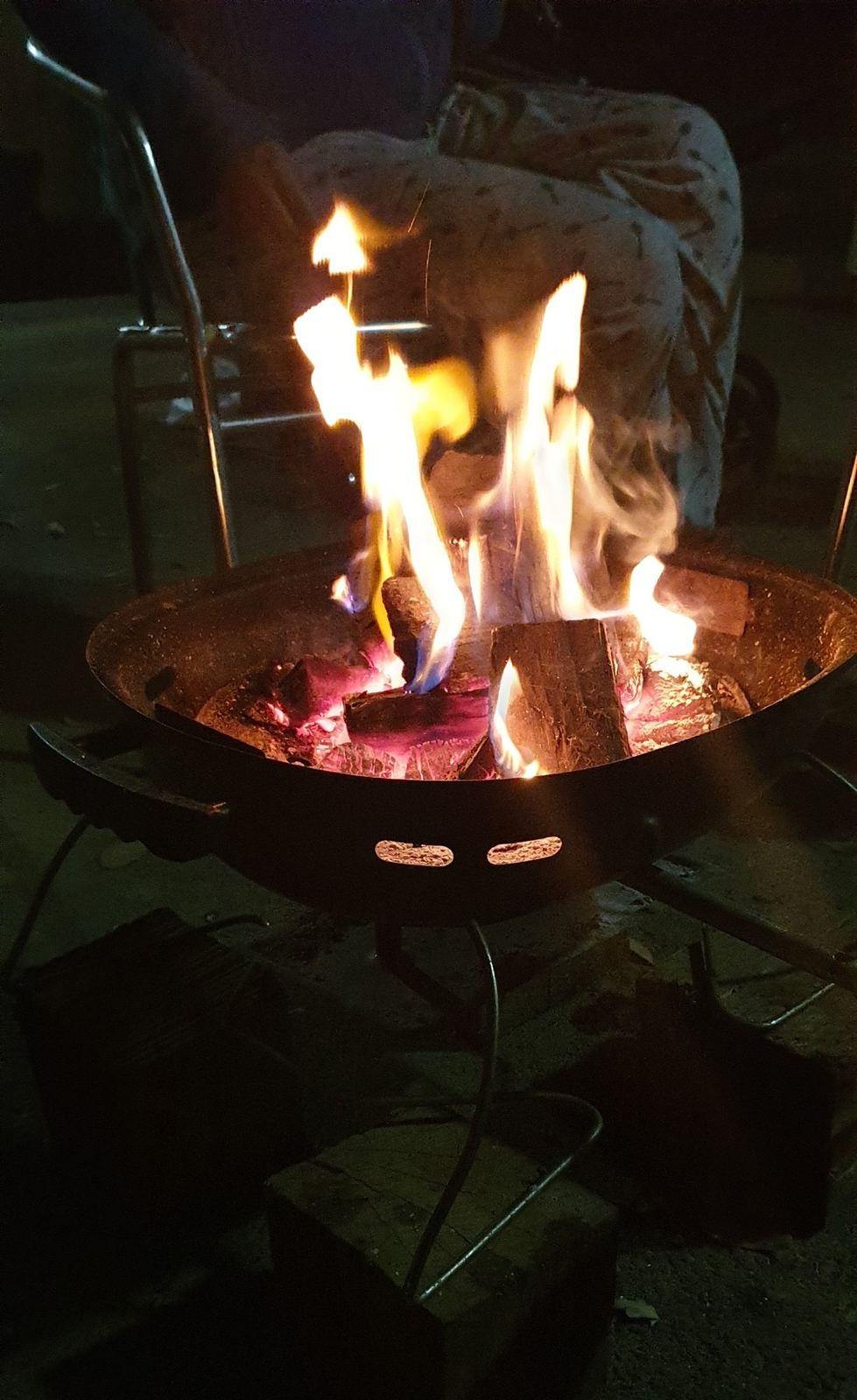 A steel fire pit bowl on legs with flames rising from split logs, glowing coals underneath, at night