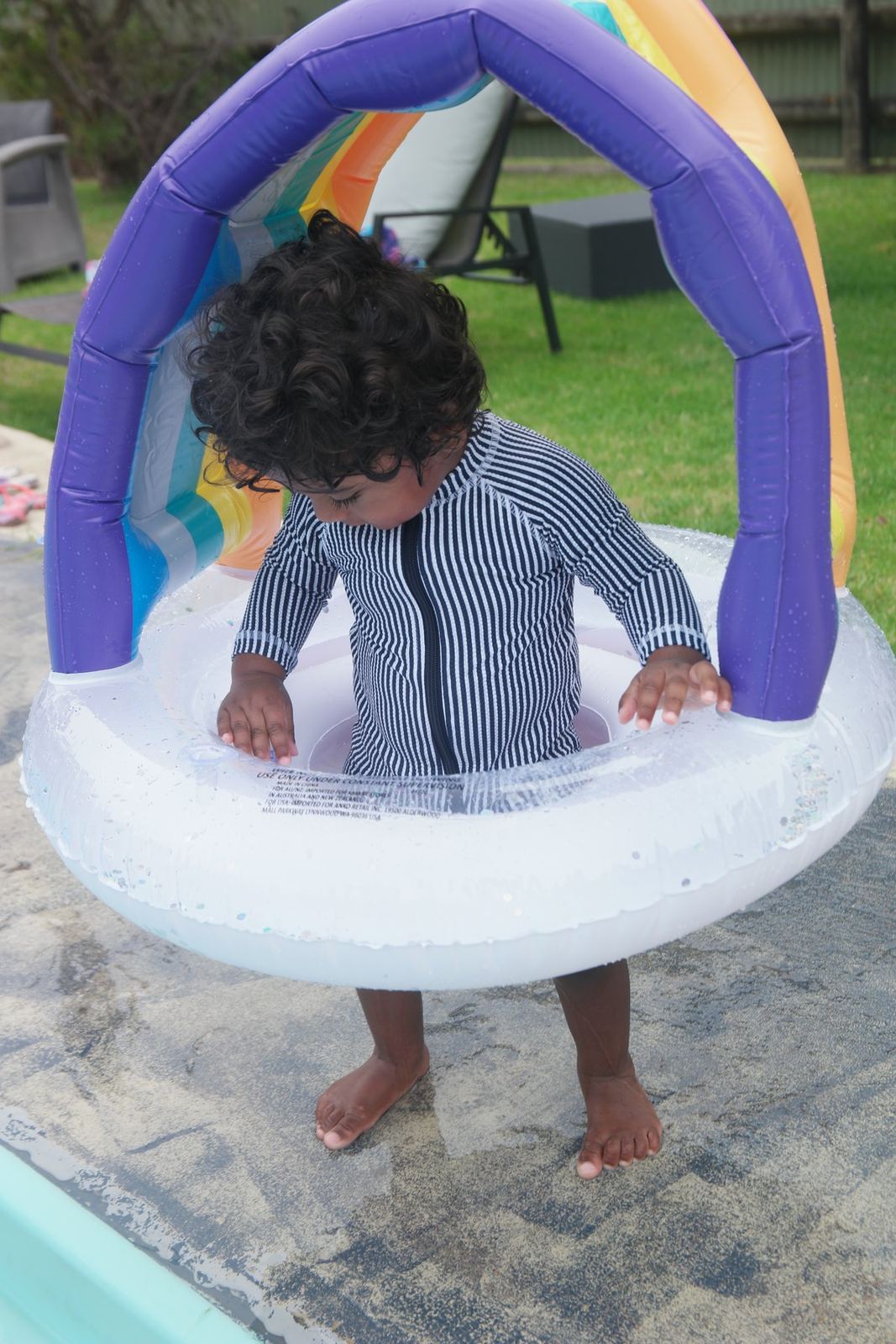 A toddler in a striped rash vest standing inside an inflatable ring pool with a rainbow canopy, on paving beside the main pool, grass and trees behind