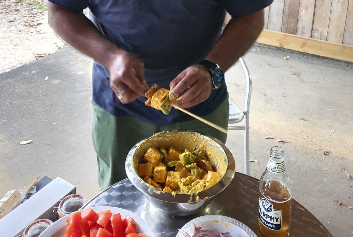 A man pulling grilled paneer and vegetables off a skewer into a large steel bowl already full of them, watermelon and drinks on the table beside