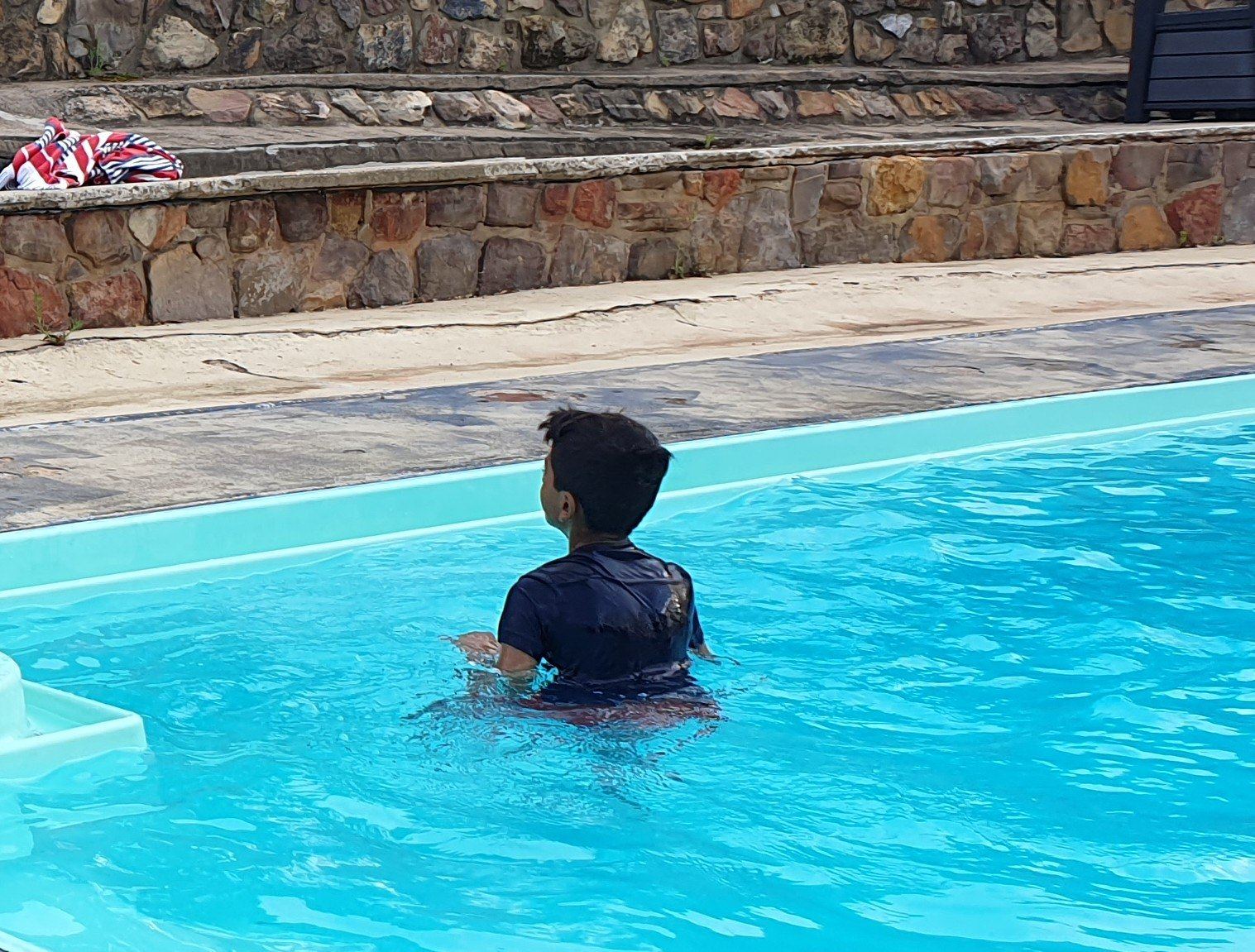 A boy standing chest-deep in a bright blue in-ground swimming pool, a stone retaining wall and a building behind
