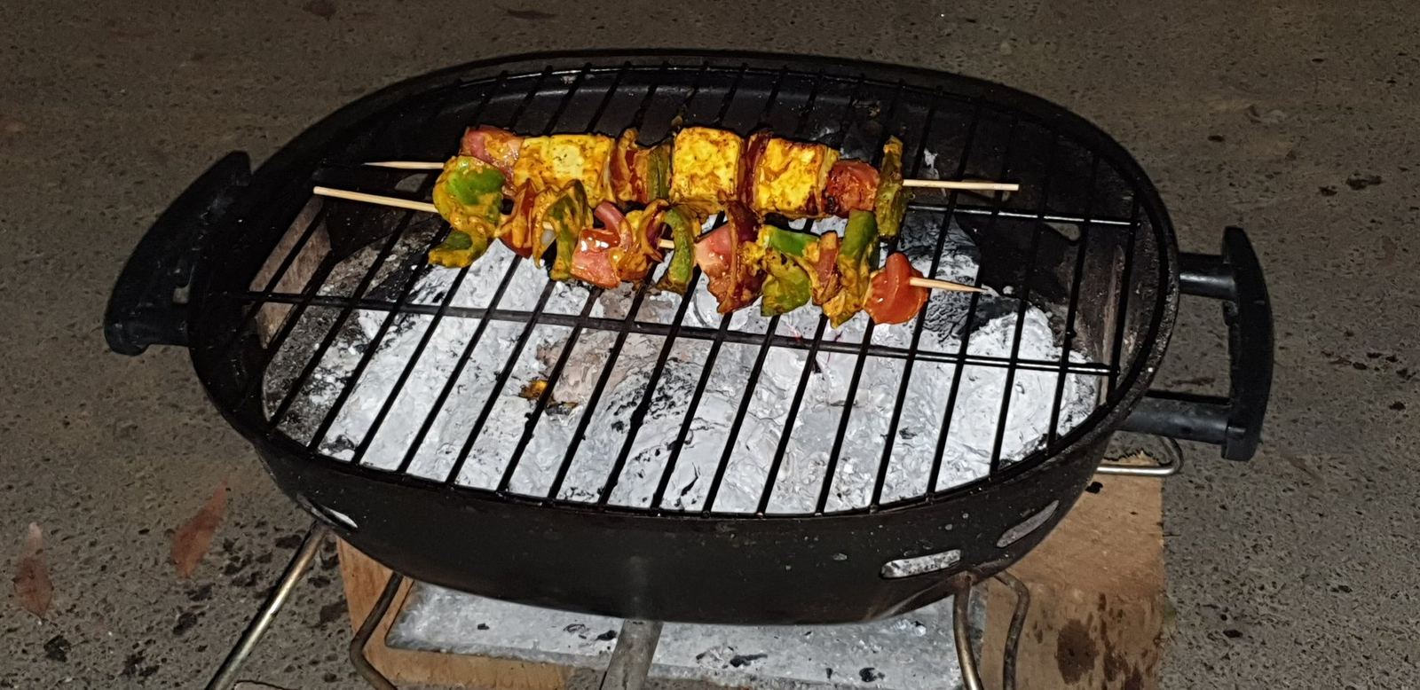 A round charcoal kettle barbecue open on a stand, a row of skewers loaded with paneer capsicum and onion laid across the grill