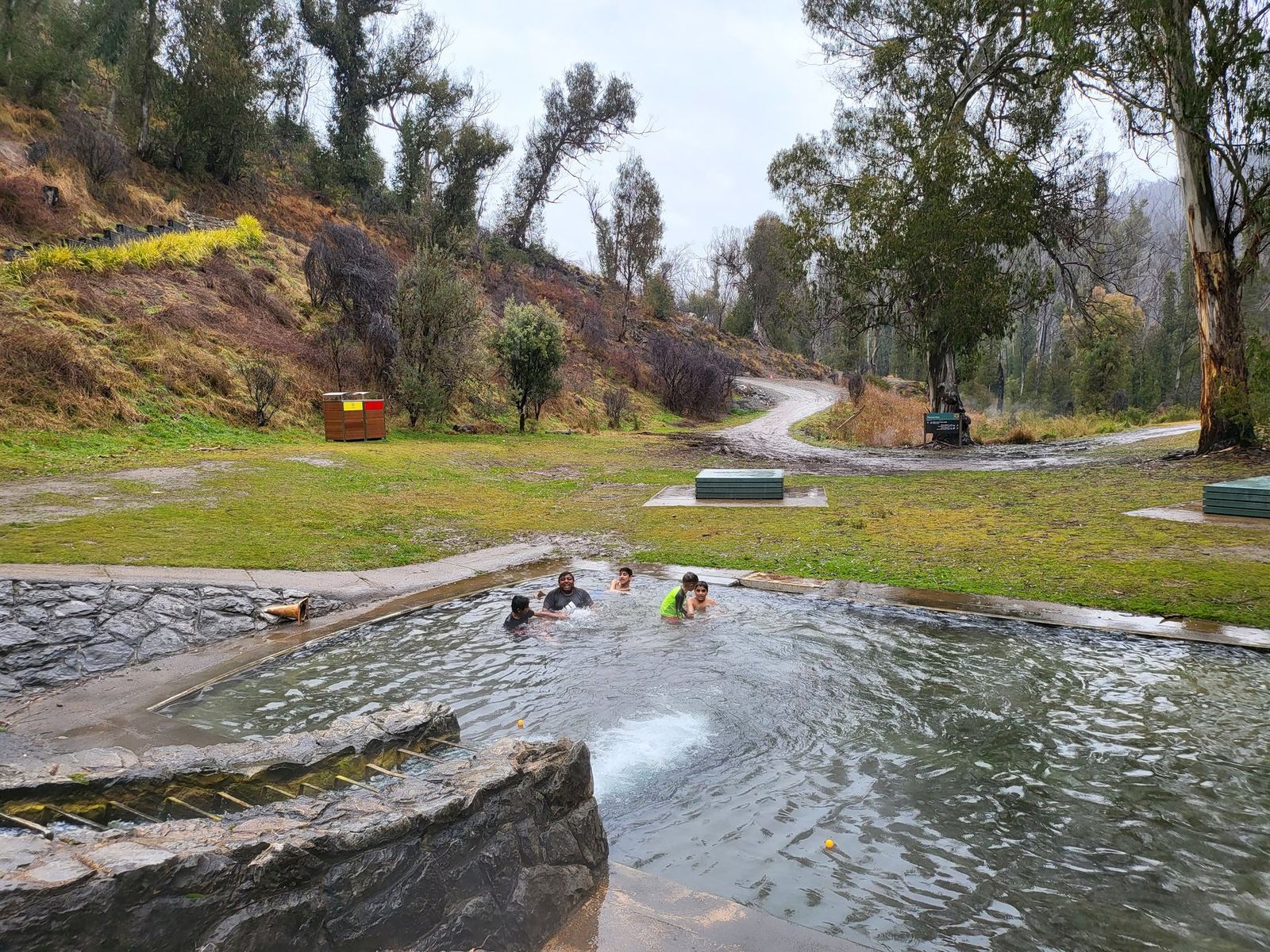 A stone-edged outdoor pool with steam rising from the surface, several people swimming in it, a grassy slope and bare winter trees rising behind and a gravel path curving away